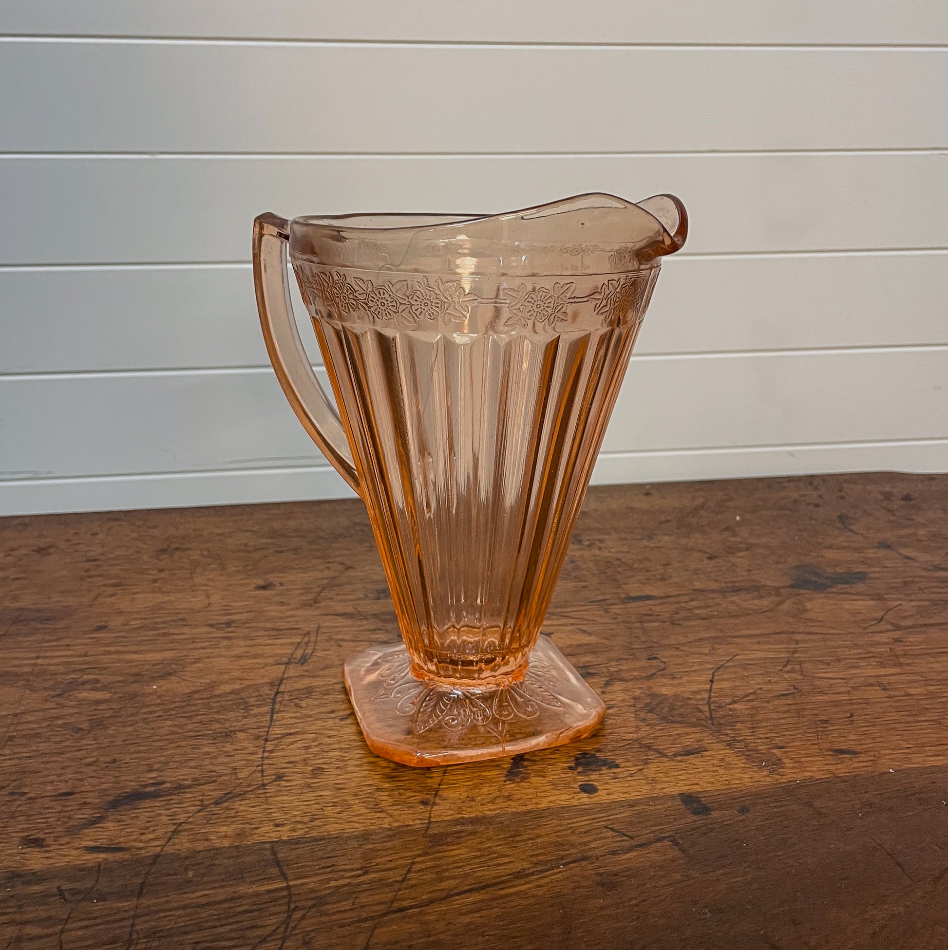 Vintage pink glass pitcher on a wooden surface with a white paneled background