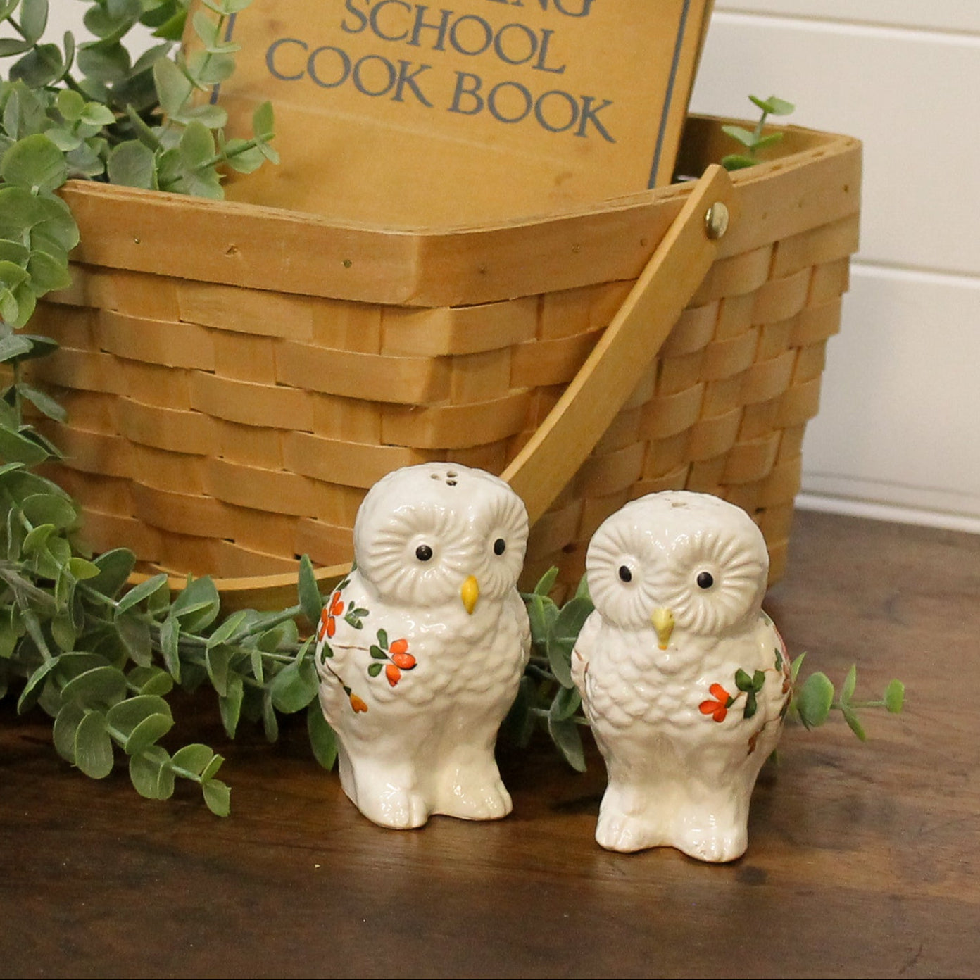 Two ceramic owl figurines with a wooden basket and cook book in the background.