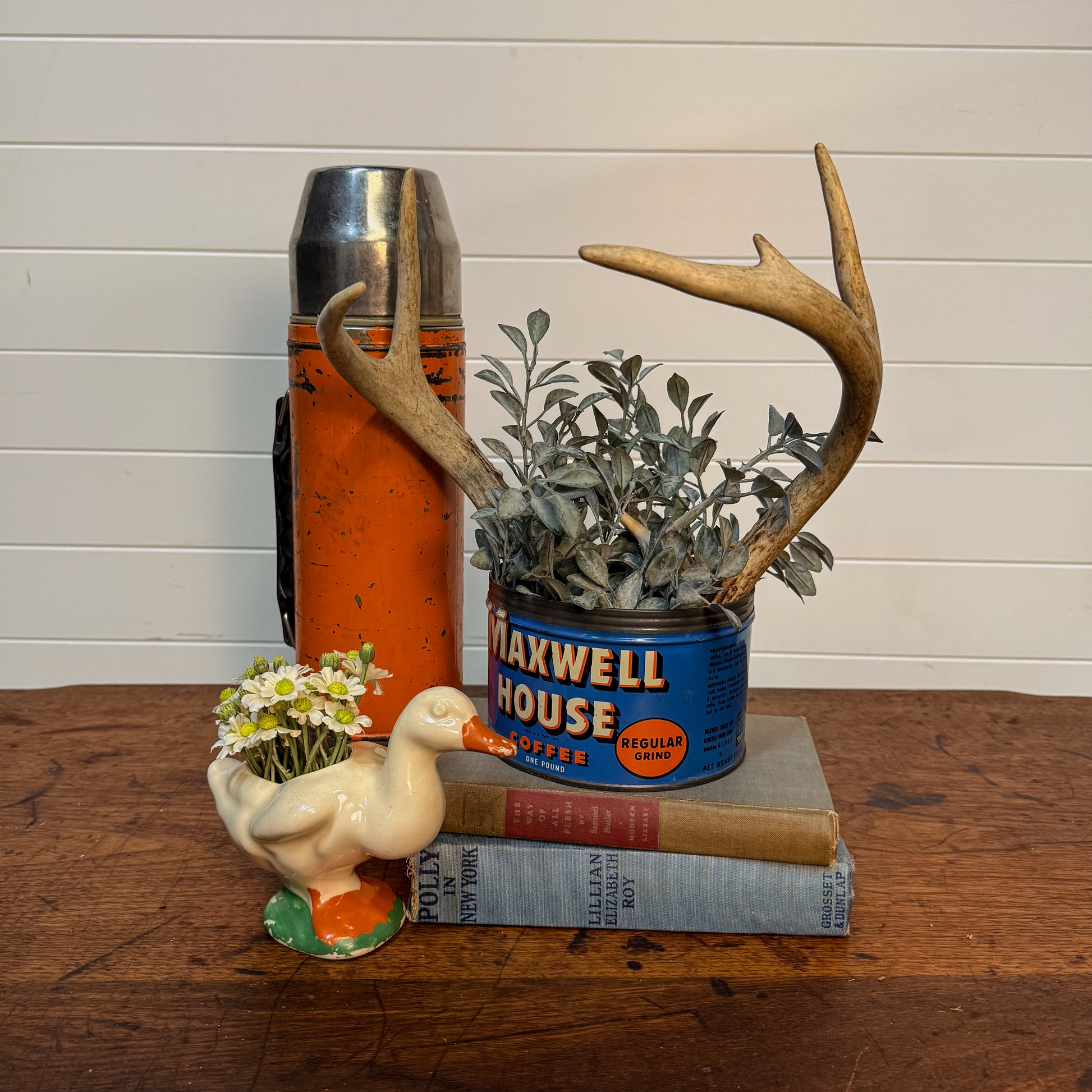 Decorative items including a vintage thermos, Maxwell House coffee can, plant with antlers, and small figurine on a wooden surface.
