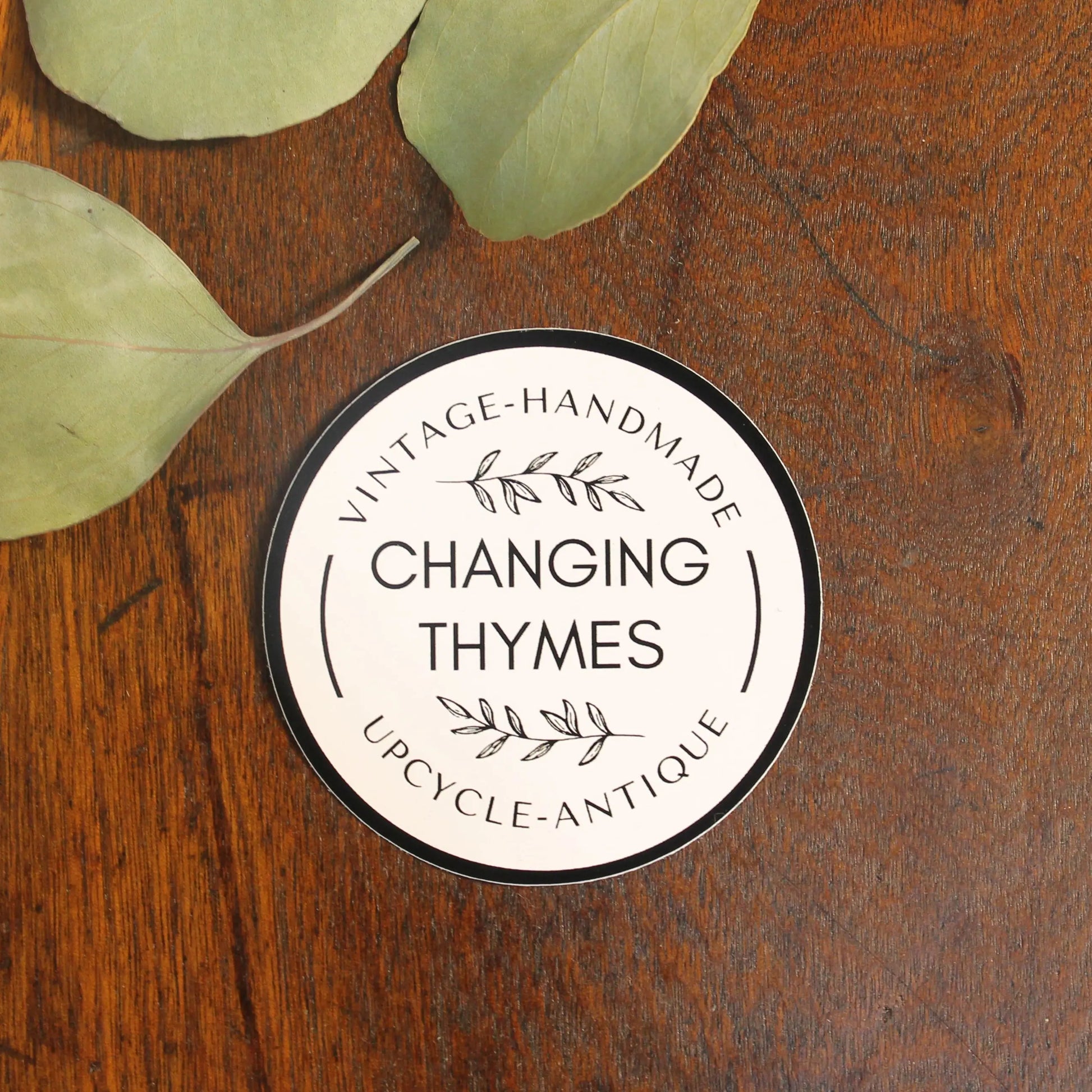 Round product label with 'Changing Thymes' on a wooden surface with leaves