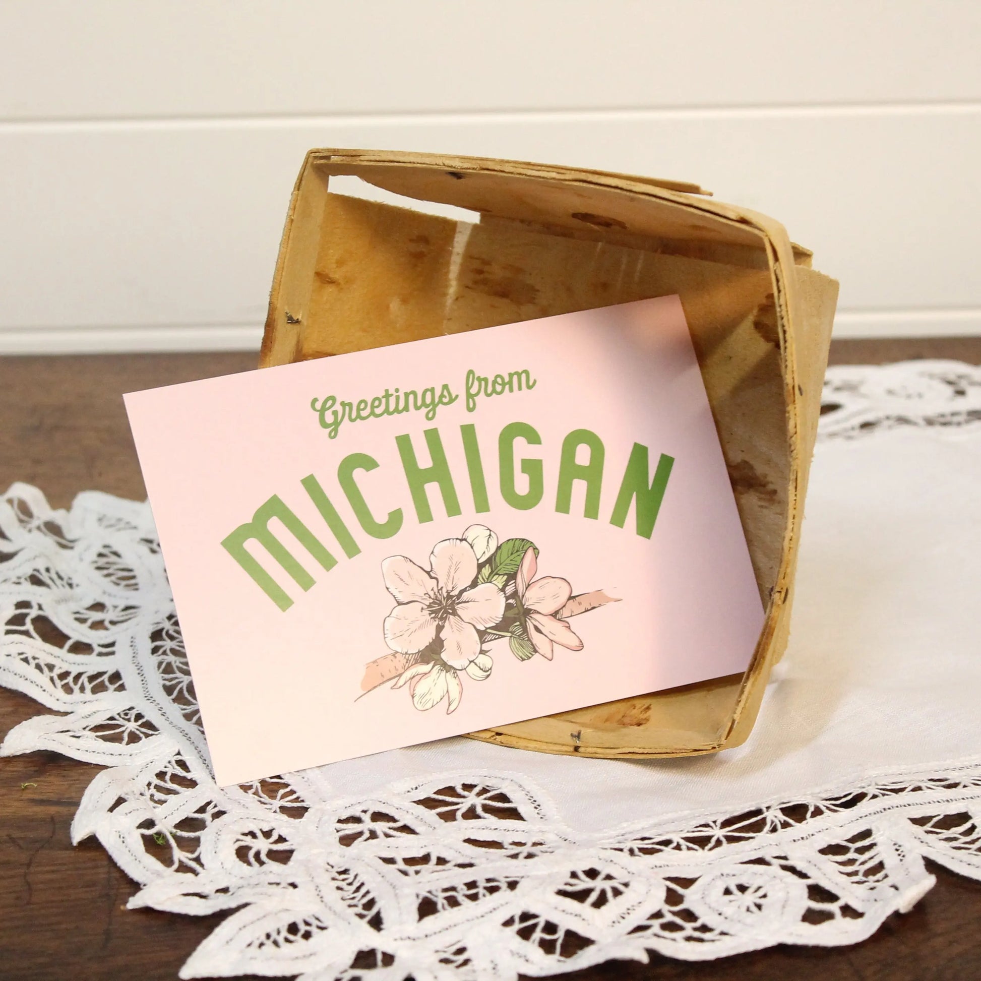 Card with 'Greetings from Michigan' text and floral design in a wooden crate on a lace doily.