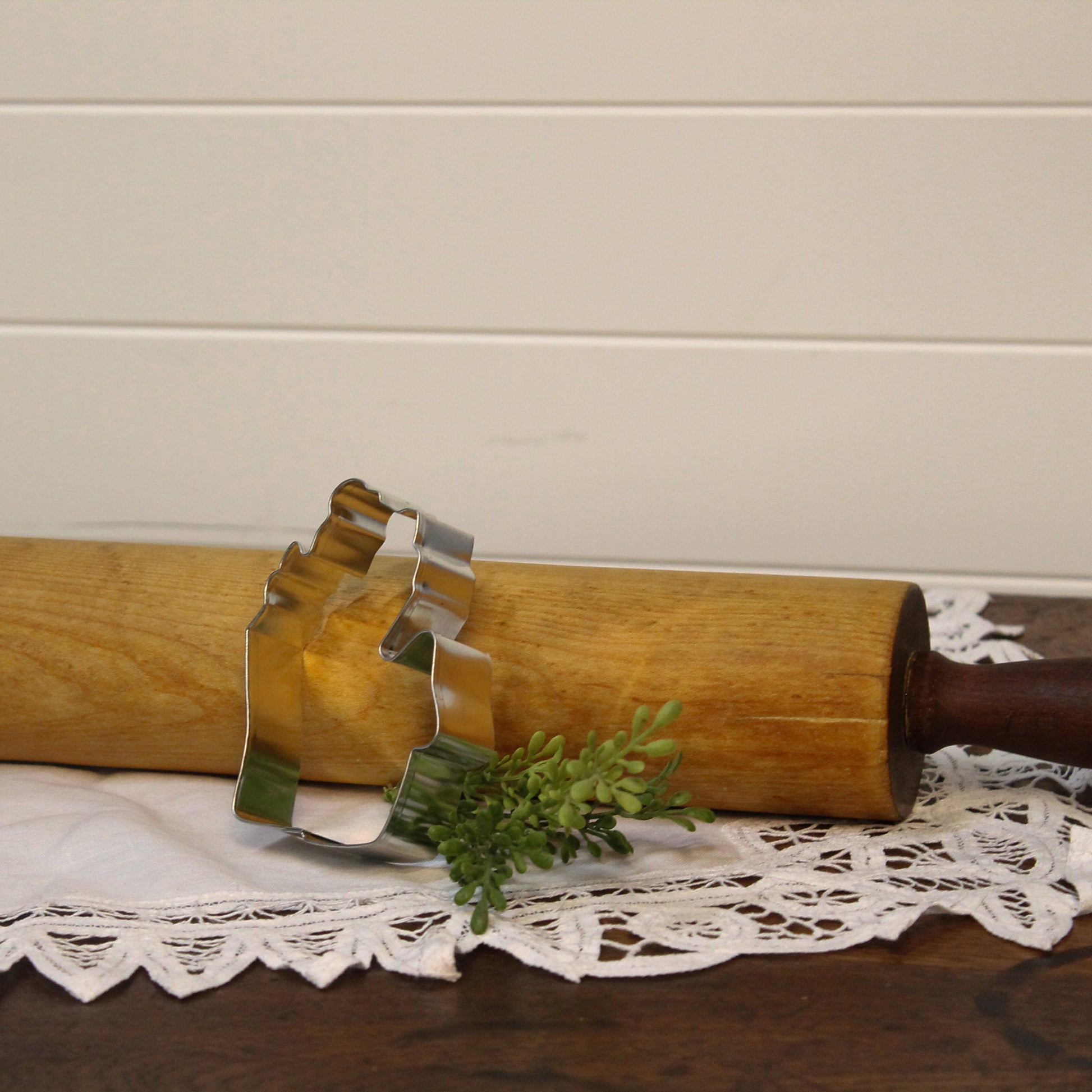 Wooden rolling pin with metal cookie cutters on a lace tablecloth against a beige wall.