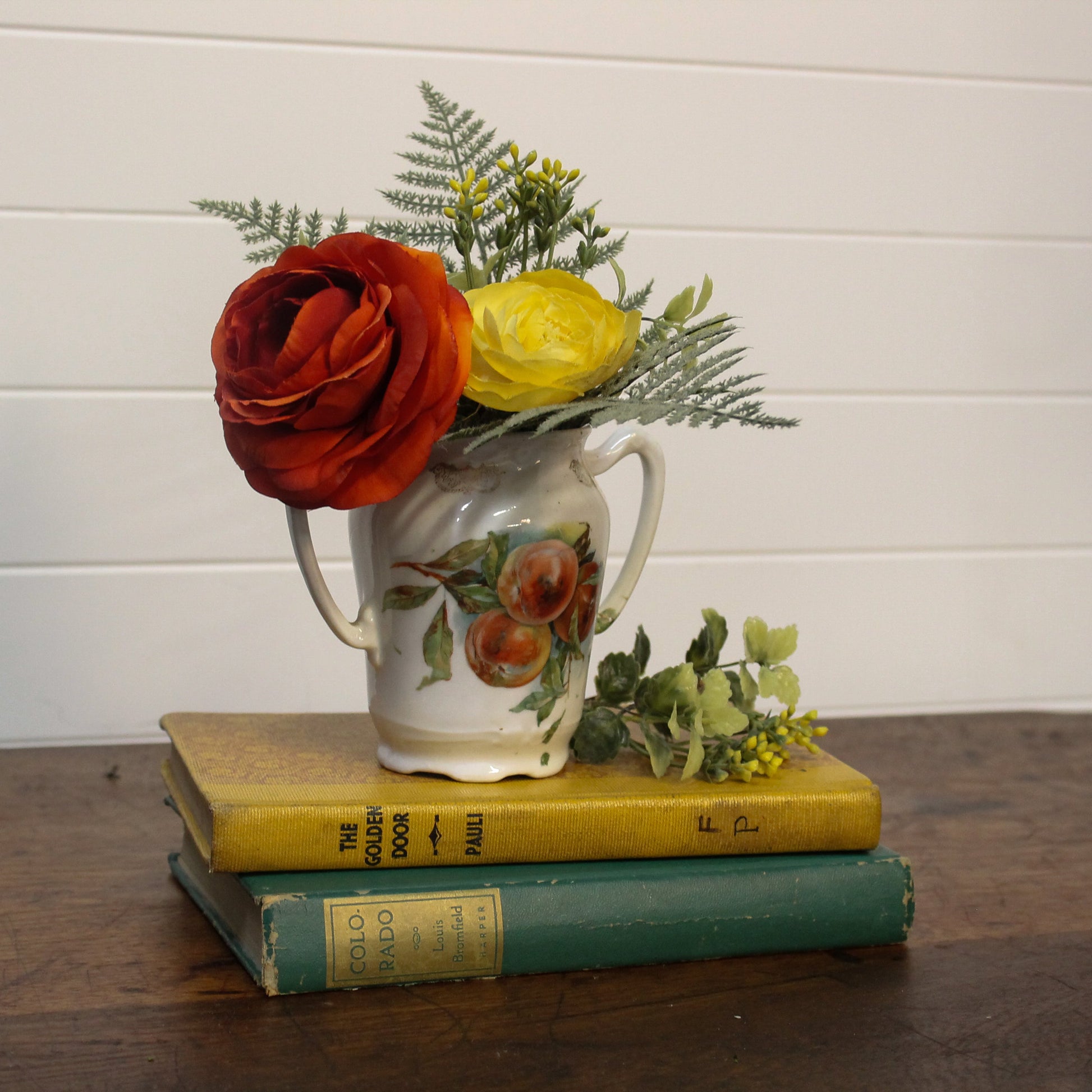Floral arrangement in a ceramic pitcher on top of two books with a white wooden panel background