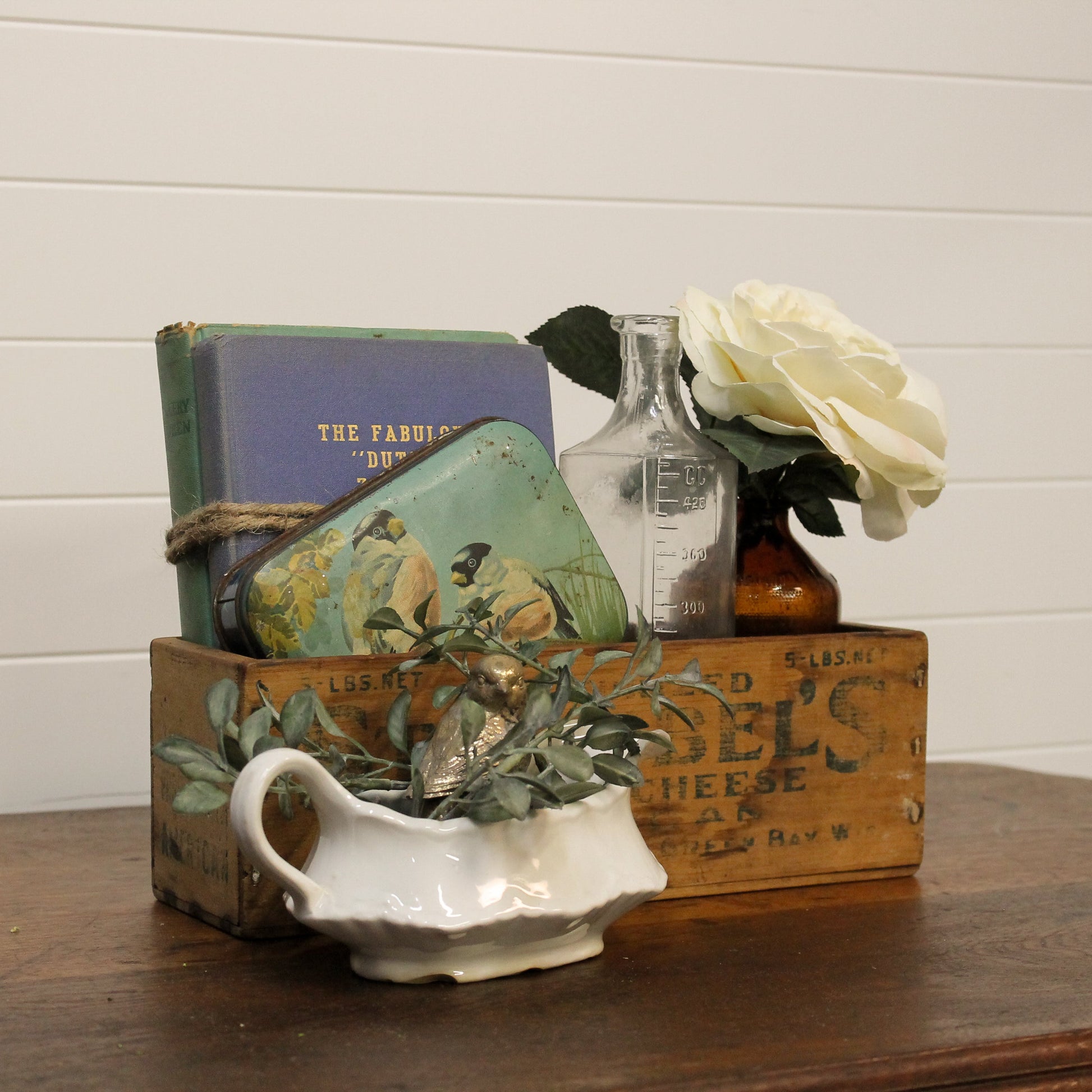 Decorative arrangement with vintage books, a bottle, a white rose, and a small pitcher on a wooden surface.