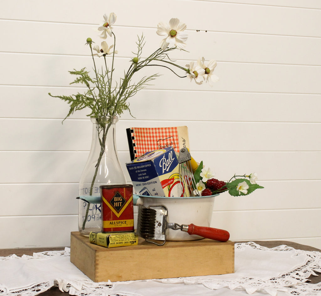 Vintage kitchen items including a can opener, flour sifter, and a bottle with flowers on a wooden block against a white wooden panel background.