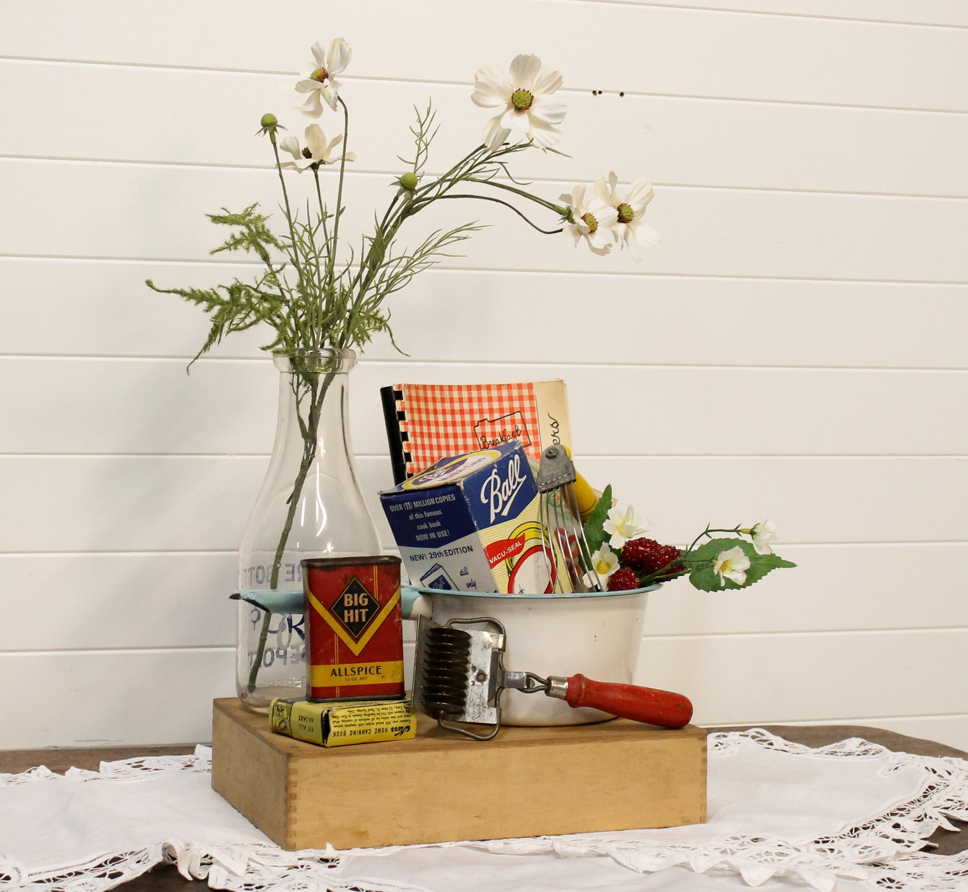 Vintage kitchen items including a can opener, flour sifter, and a bottle with flowers on a wooden block against a white wooden panel background.