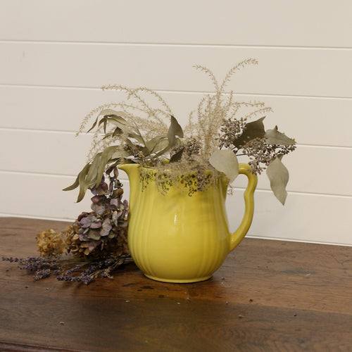 Yellow pitcher with decorative plants on a wooden surface against a white wall