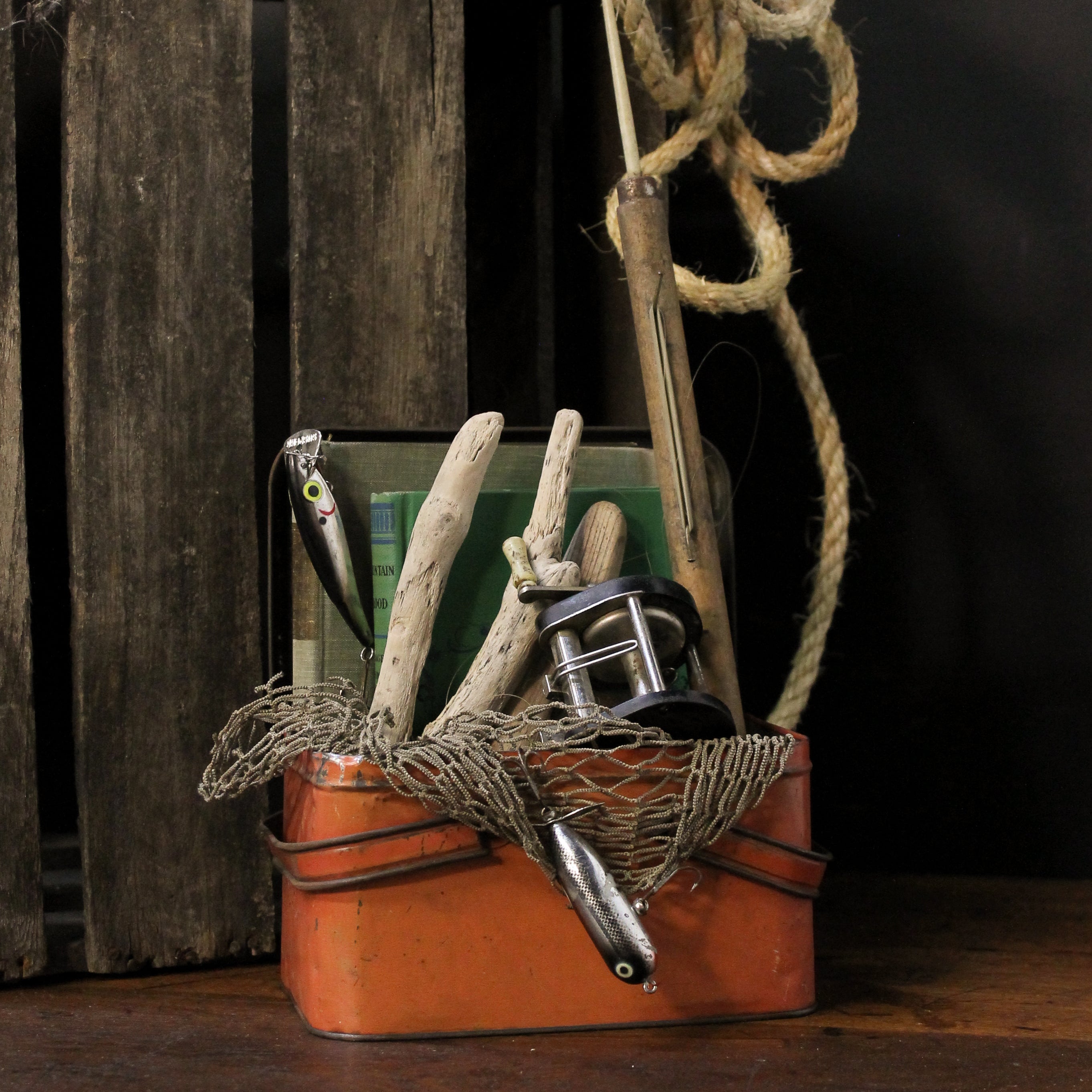 Decorative arrangement with fishing reel, lures, and driftwood in a leather basket against a wooden crate background.