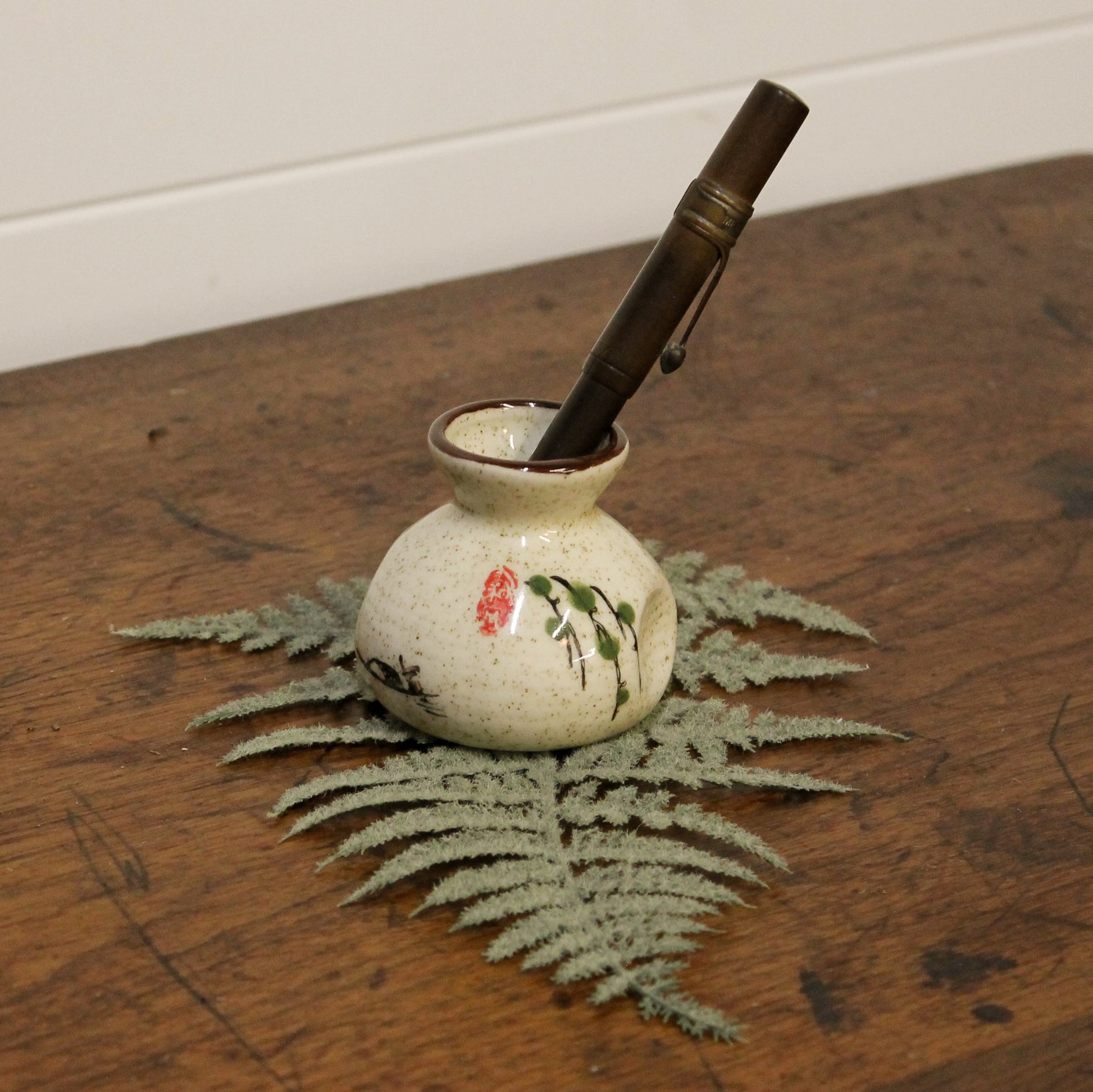 Small ceramic pot with a pen on a wooden surface with a fern leaf