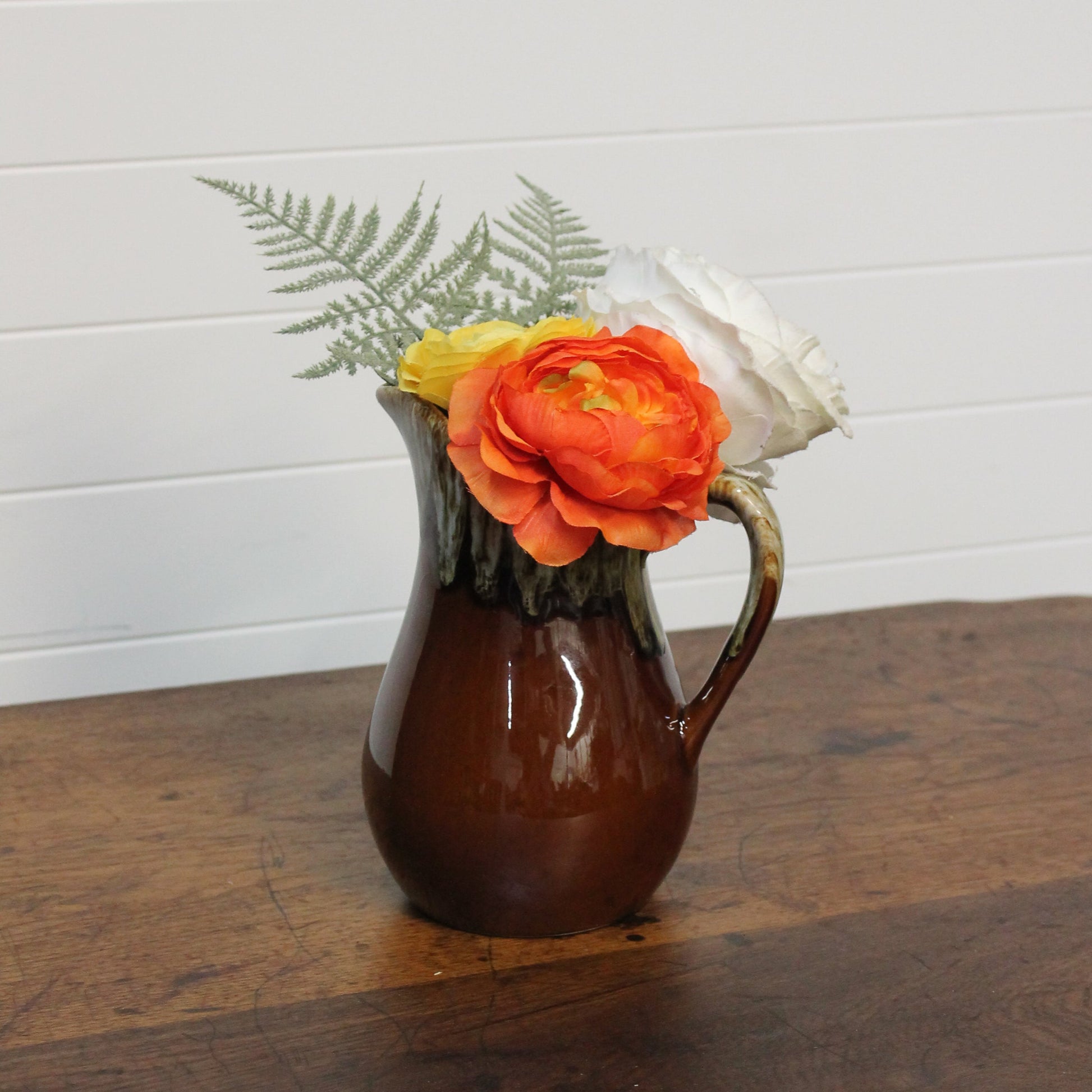 Brown pitcher with flowers on a wooden surface against a white wall