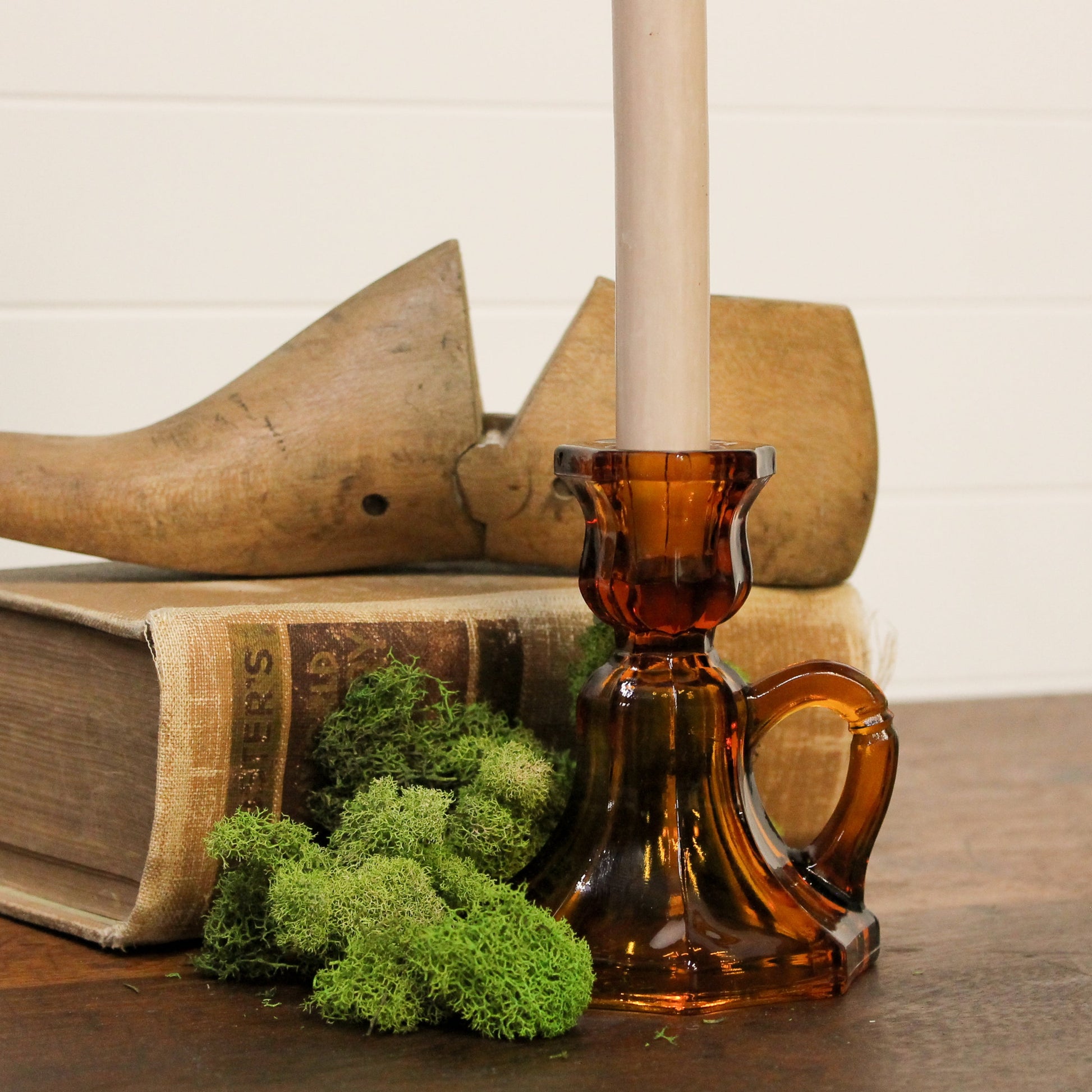 Brown glass candle holder with a candle, book, wooden shoe, and greenery on a wooden surface.
