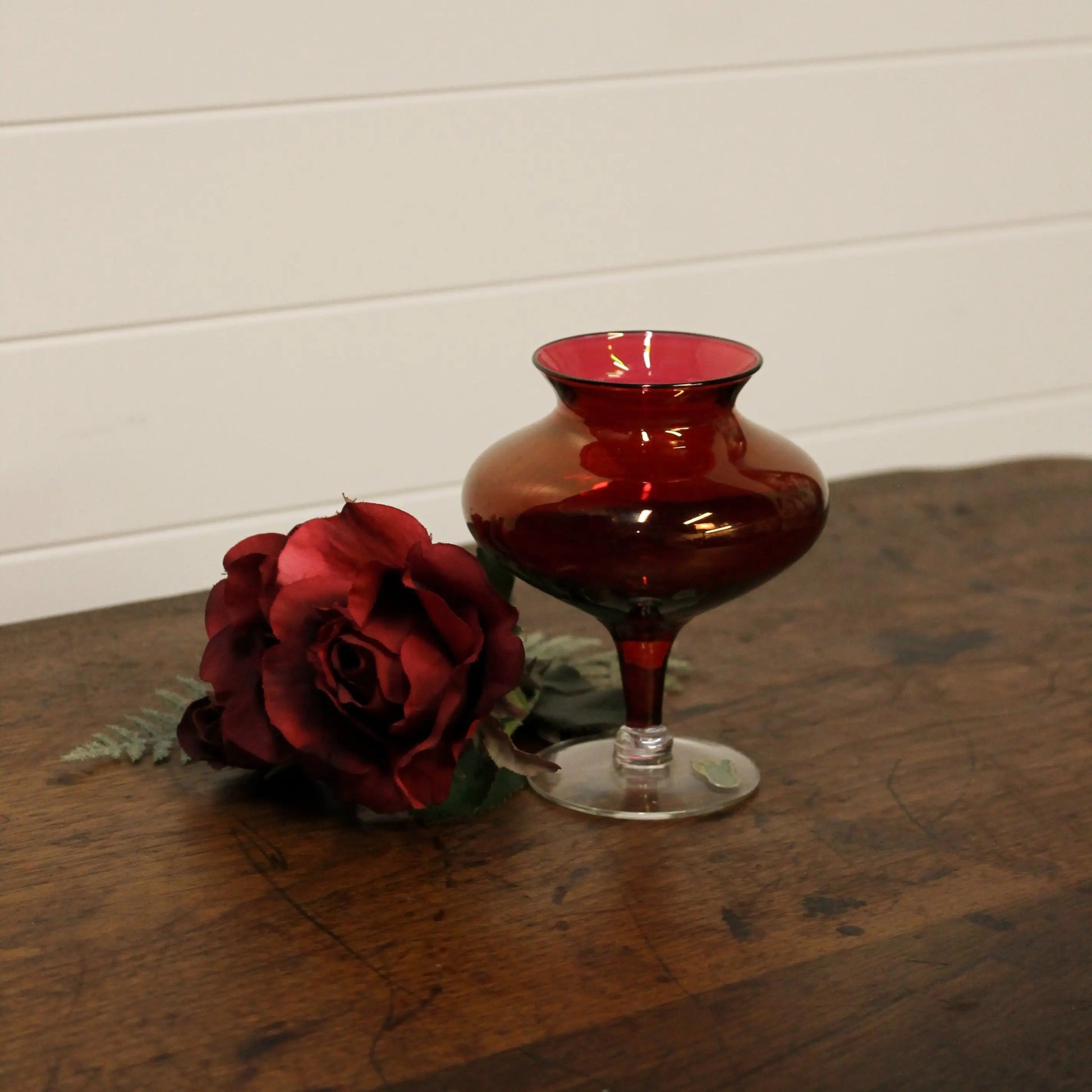 Red glass vase and red rose on a wooden surface with a white wall background