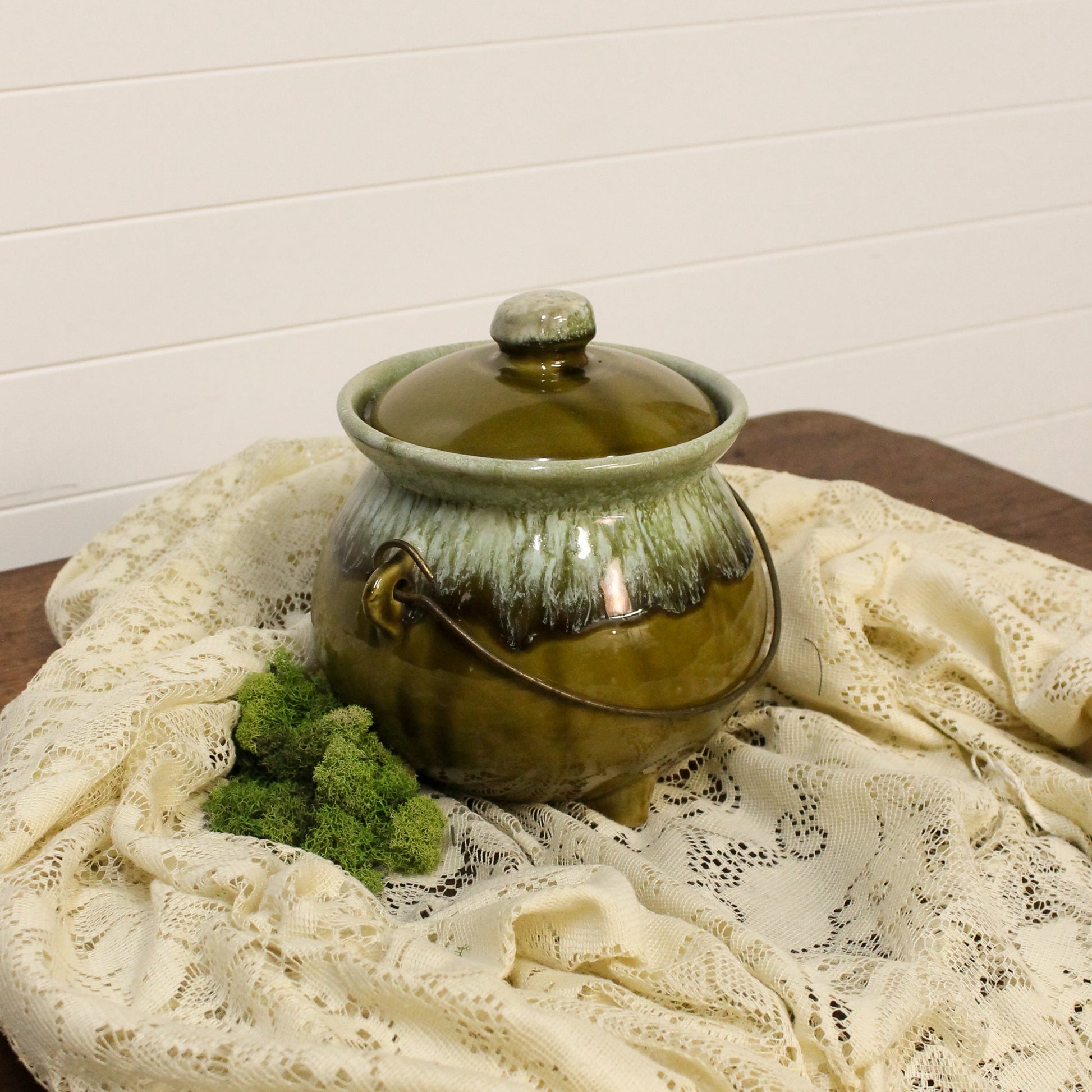 Green ceramic pot with a lid on a lace tablecloth against a white wall.